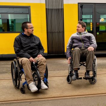 Zwei Menschen im Rollstuhl unterhalten sich vor einer Straßenbahn der BVG