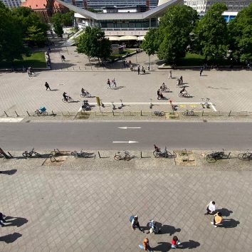 Blick aus der Vogelperspektive auf eine Fußgängerzone und einen Radweg nahe des Berliner Fernsehturms am Alexanderplatz. Diverse Menschen bewegen sich in alle Richtungen zu Fuß, per Rad oder mit Kinderwagen.