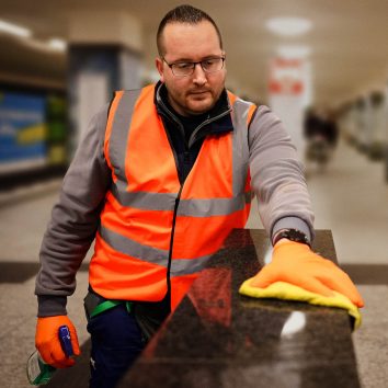 Eine Reinigungskraft säubert mit einem Lappen eine Oberfläche und hält in der anderen Hand das Reinigungsmittel. Die Person befindet sich in einem U-Bahnhof. Im Hintergrund ist eine U-Bahn zu erkennen.