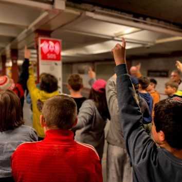 Teilnehmende Schulkinder der Verkehrserziehung heben die Hand, um eine Frage zu beantworten. Sie stehen vor der Informationssäule auf einem Bahnsteig.